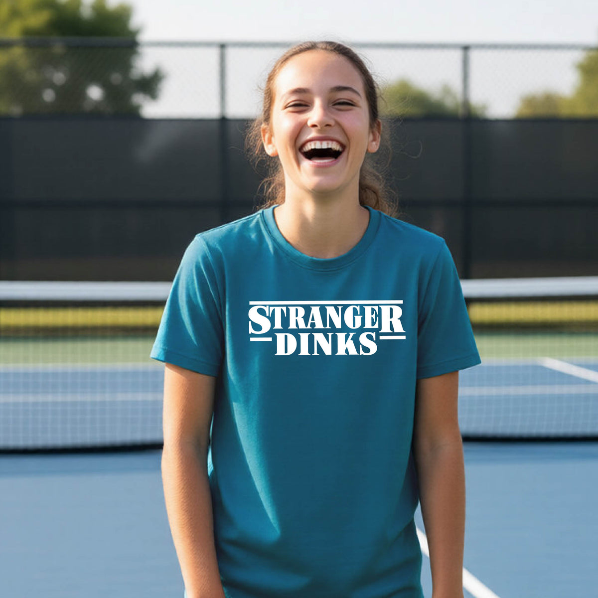 A young person on a pickleball court wearing a teal short-sleeve t-shirt with a white graphic of the words “STRANGER DINKS” on the front of the shirt in the style of the popular TV Series Stranger Things. 