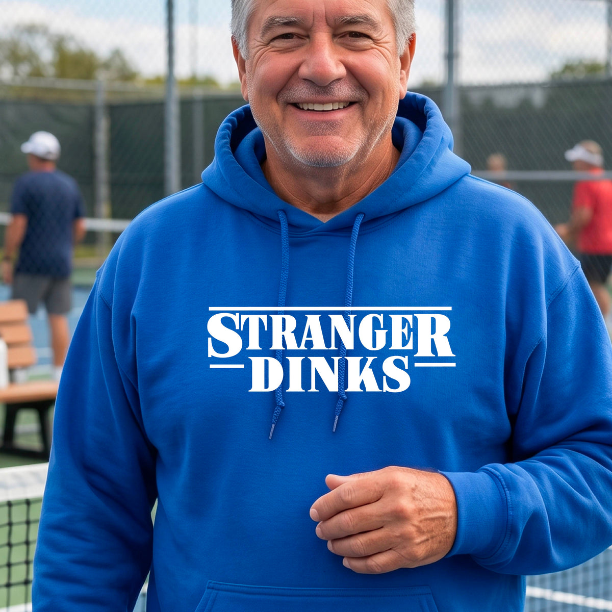 A man on a pickleball court wearing a royal blue hoodie pullover with a white graphic of the words “STRANGER DINKS” on the front of the shirt in the style of the popular TV Series Stranger Things. 