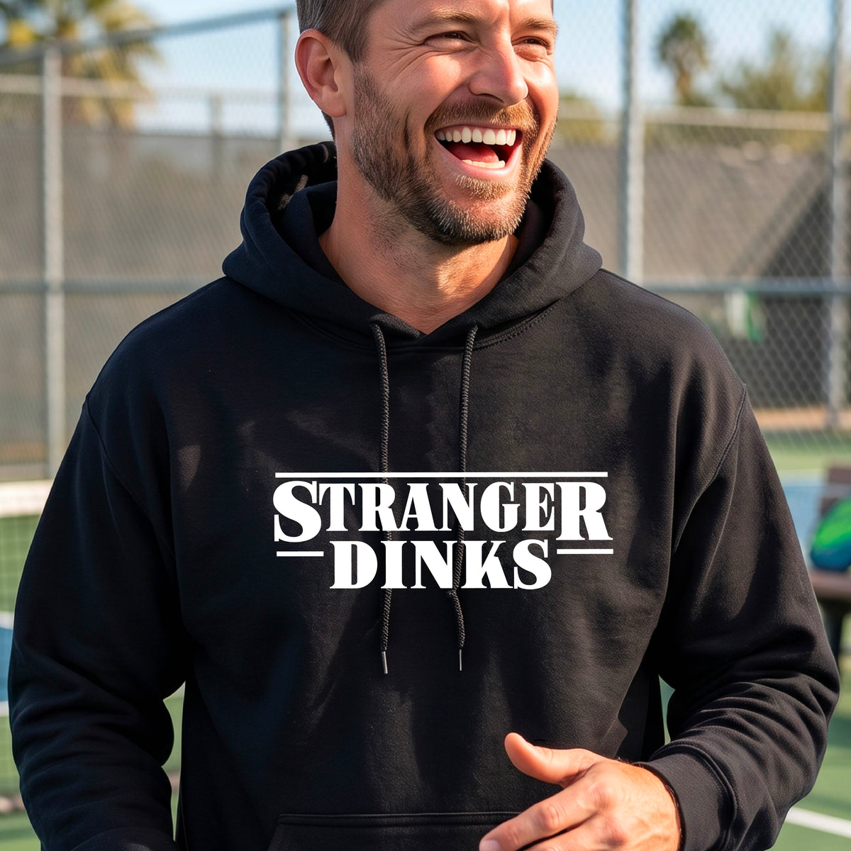 A man on a pickleball court wearing a black hoodie pullover with a white graphic of the words “STRANGER DINKS” on the front of the shirt in the style of the popular TV Series Stranger Things. 