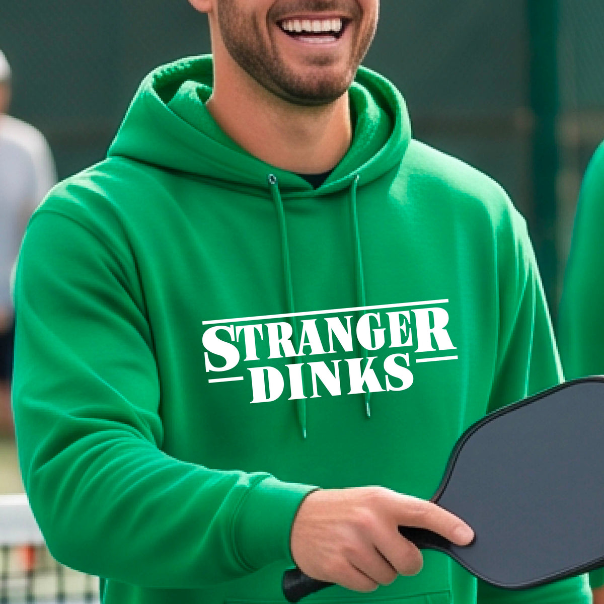 A man on a pickleball court wearing a green hoodie pullover with a white graphic of the words “STRANGER DINKS” on the front of the shirt in the style of the popular TV Series Stranger Things. 