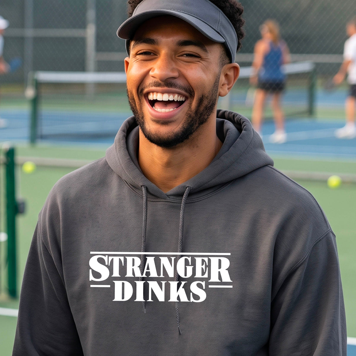 A man on a pickleball court wearing a gray hoodie pullover with a white graphic of the words “STRANGER DINKS” on the front of the shirt in the style of the popular TV Series Stranger Things. 