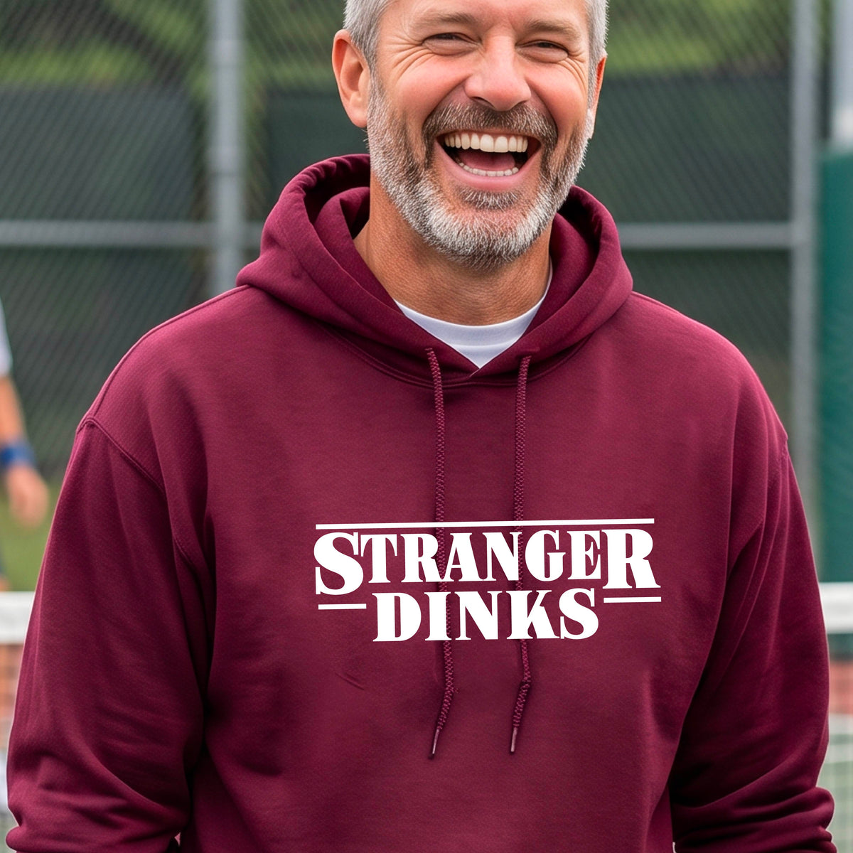 A man on a pickleball court wearing a  maroon hoodie pullover with a white graphic of the words “STRANGER DINKS” on the front of the shirt in the style of the popular TV Series Stranger Things. 