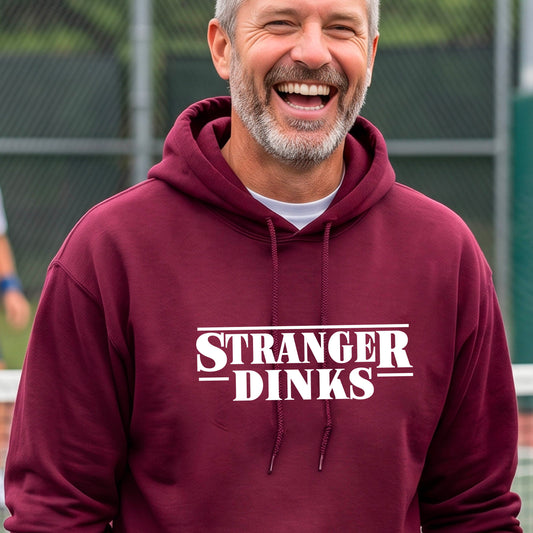 A man on a pickleball court wearing a  maroon hoodie pullover with a white graphic of the words “STRANGER DINKS” on the front of the shirt in the style of the popular TV Series Stranger Things. 