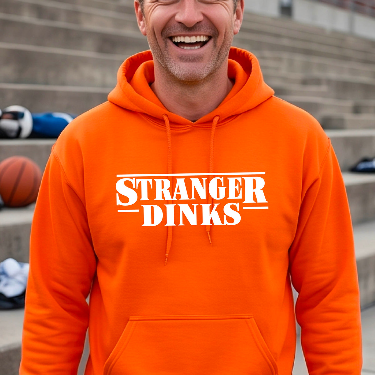 A man on a pickleball court wearing an orange hoodie pullover with a white graphic of the words “STRANGER DINKS” on the front of the shirt in the style of the popular TV Series Stranger Things. 