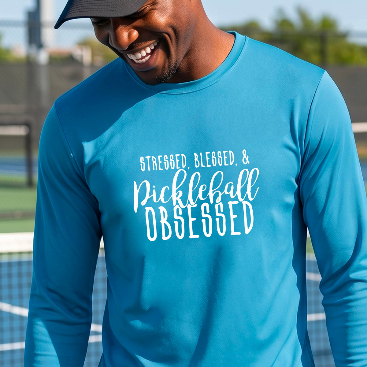 A man on a pickleball court wearing a bright blue long-sleeve t-shirt with a white graphic of the words “Stressed, Blessed & Pickleball Obsessed” on the front of the shirt.