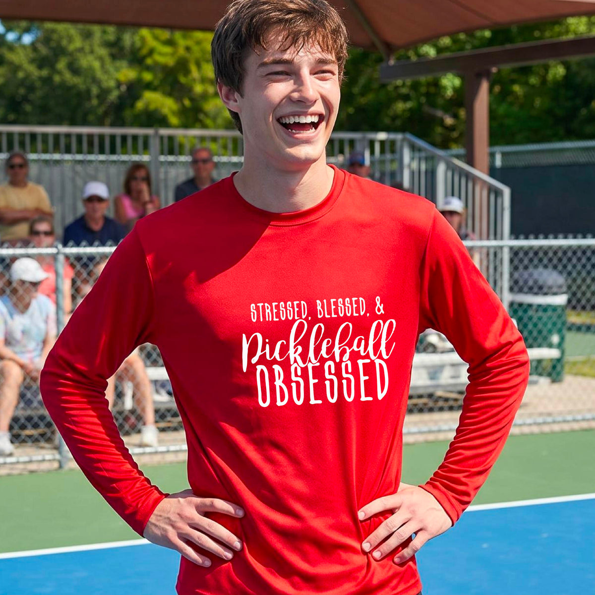 A man on a pickleball court wearing a red long-sleeve t-shirt with a white graphic of the words “Stressed, Blessed & Pickleball Obsessed” on the front of the shirt.