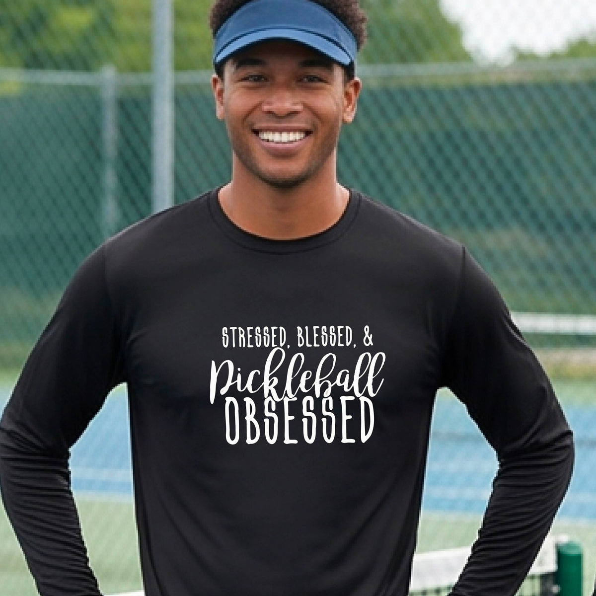 A man on a pickleball court wearing a black long-sleeve t-shirt with a white graphic of the words “Stressed, Blessed & Pickleball Obsessed” on the front of the shirt.