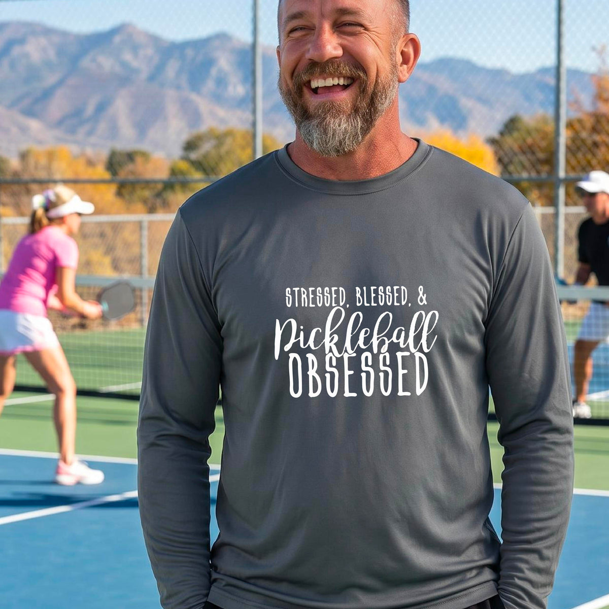 A man on a pickleball court wearing a dark gray long-sleeve t-shirt with a white graphic of the words “Stressed, Blessed & Pickleball Obsessed” on the front of the shirt.