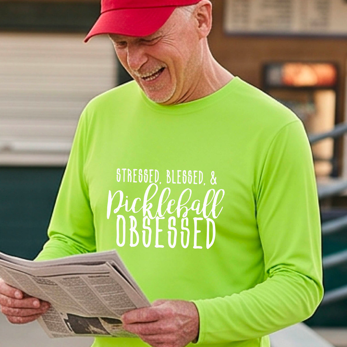 A man on a pickleball court wearing a lime green long-sleeve t-shirt with a white graphic of the words “Stressed, Blessed & Pickleball Obsessed” on the front of the shirt. 