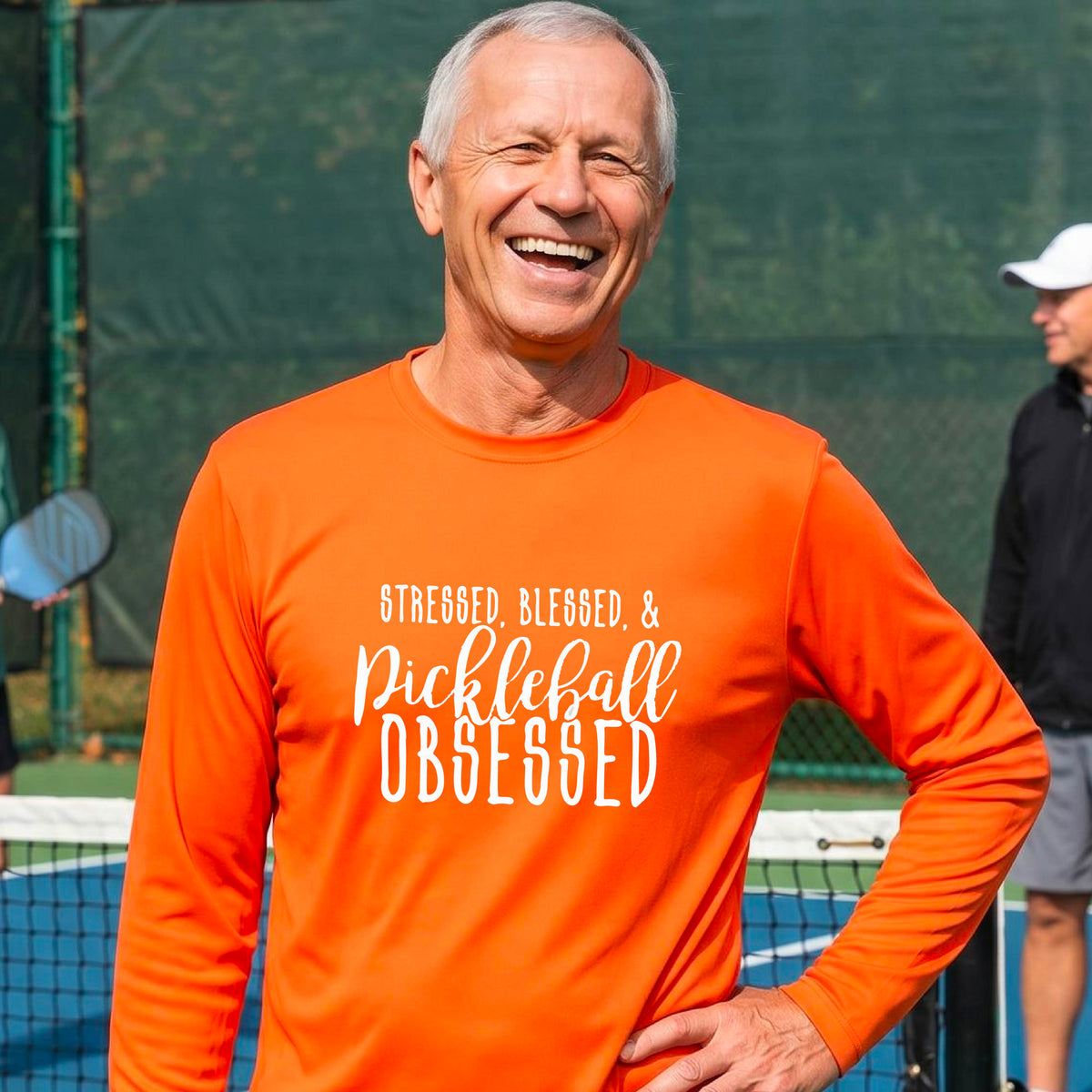 A man on a pickleball court wearing an orange long-sleeve t-shirt with a white graphic of the words “Stressed, Blessed & Pickleball Obsessed” on the front of the shirt.