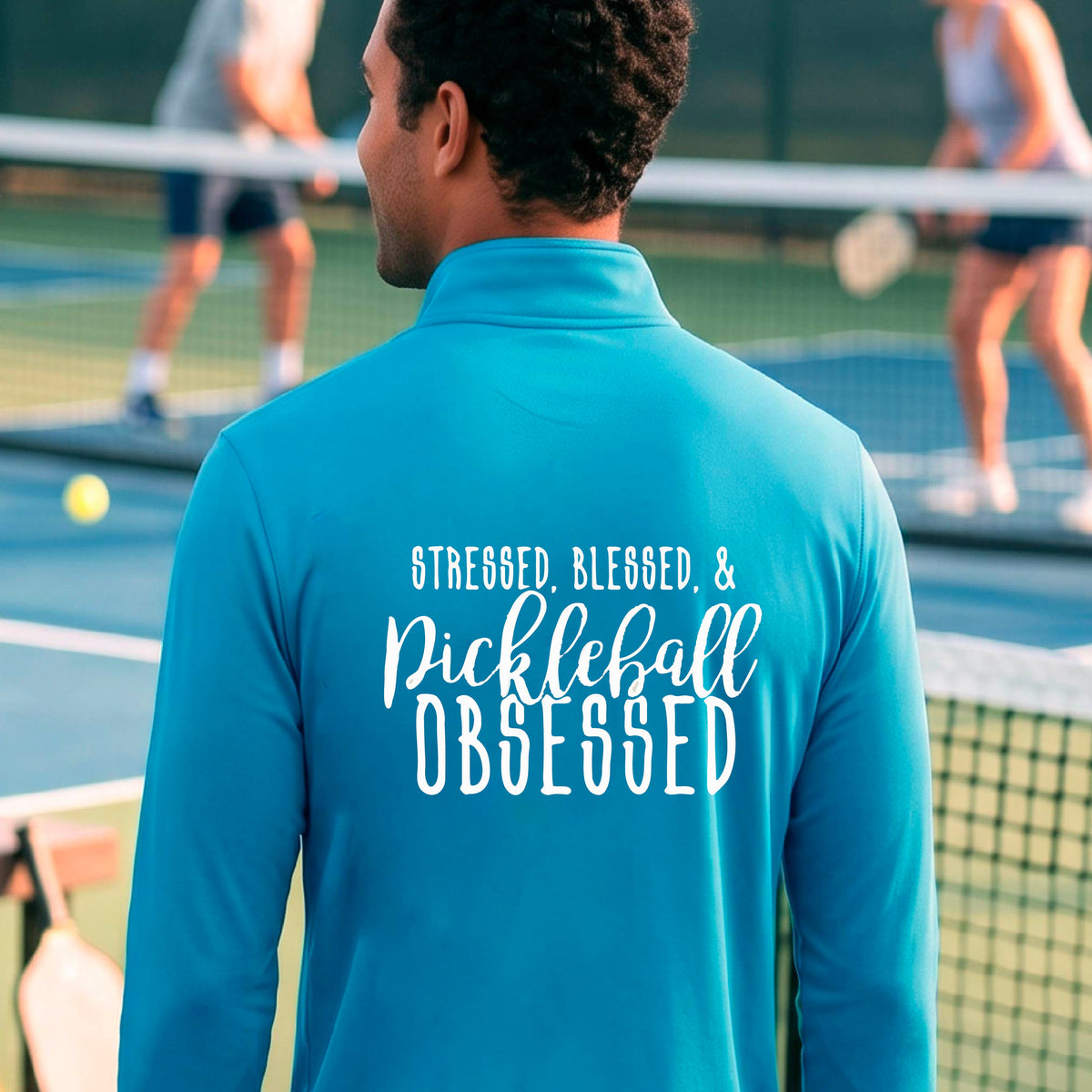 A man seen from behind on a pickleball court wearing a bright blue quarter-zip shirt with a white graphic of the words “Stressed, Blessed & Pickleball Obsessed” on the back of the shirt.