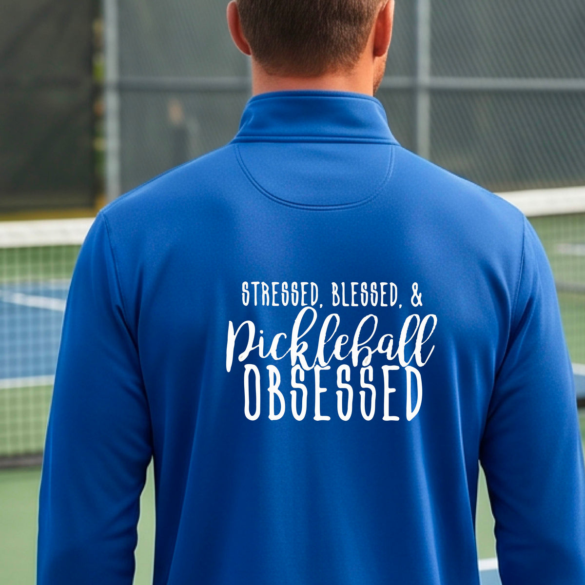 A man seen from behind on a pickleball court wearing a royal blue quarter-zip shirt with a white graphic of the words “Stressed, Blessed & Pickleball Obsessed” on the back of the shirt.