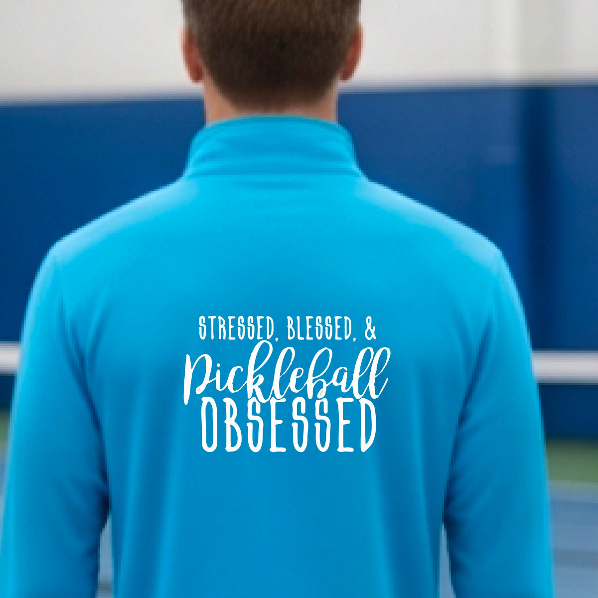 A man seen from behind on a pickleball court wearing a bright blue quarter-zip shirt with a white graphic of the words “Stressed, Blessed & Pickleball Obsessed” on the back of the shirt.
