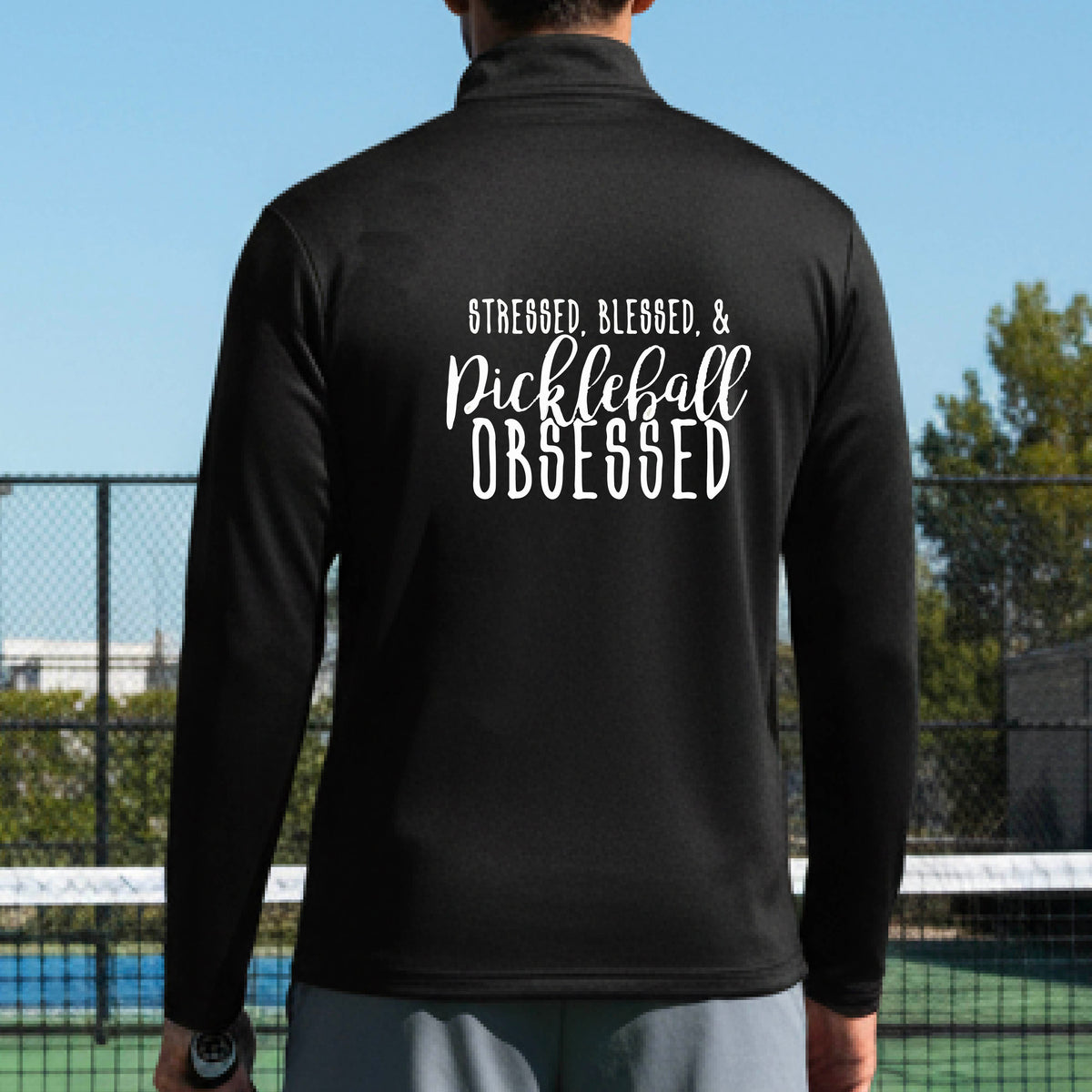 A man seen from behind on a pickleball court wearing a black quarter-zip shirt with a white graphic of the words “Stressed, Blessed & Pickleball Obsessed” on the back of the shirt.