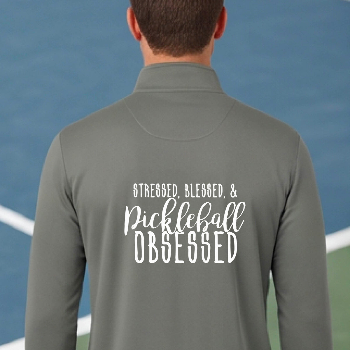 A man seen from behind on a pickleball court wearing a gray quarter-zip shirt with a white graphic of the words “Stressed, Blessed & Pickleball Obsessed” on the back of the shirt.