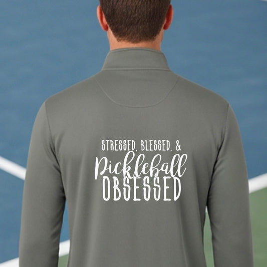 A man seen from behind on a pickleball court wearing a gray quarter-zip shirt with a white graphic of the words “Stressed, Blessed & Pickleball Obsessed” on the back of the shirt.