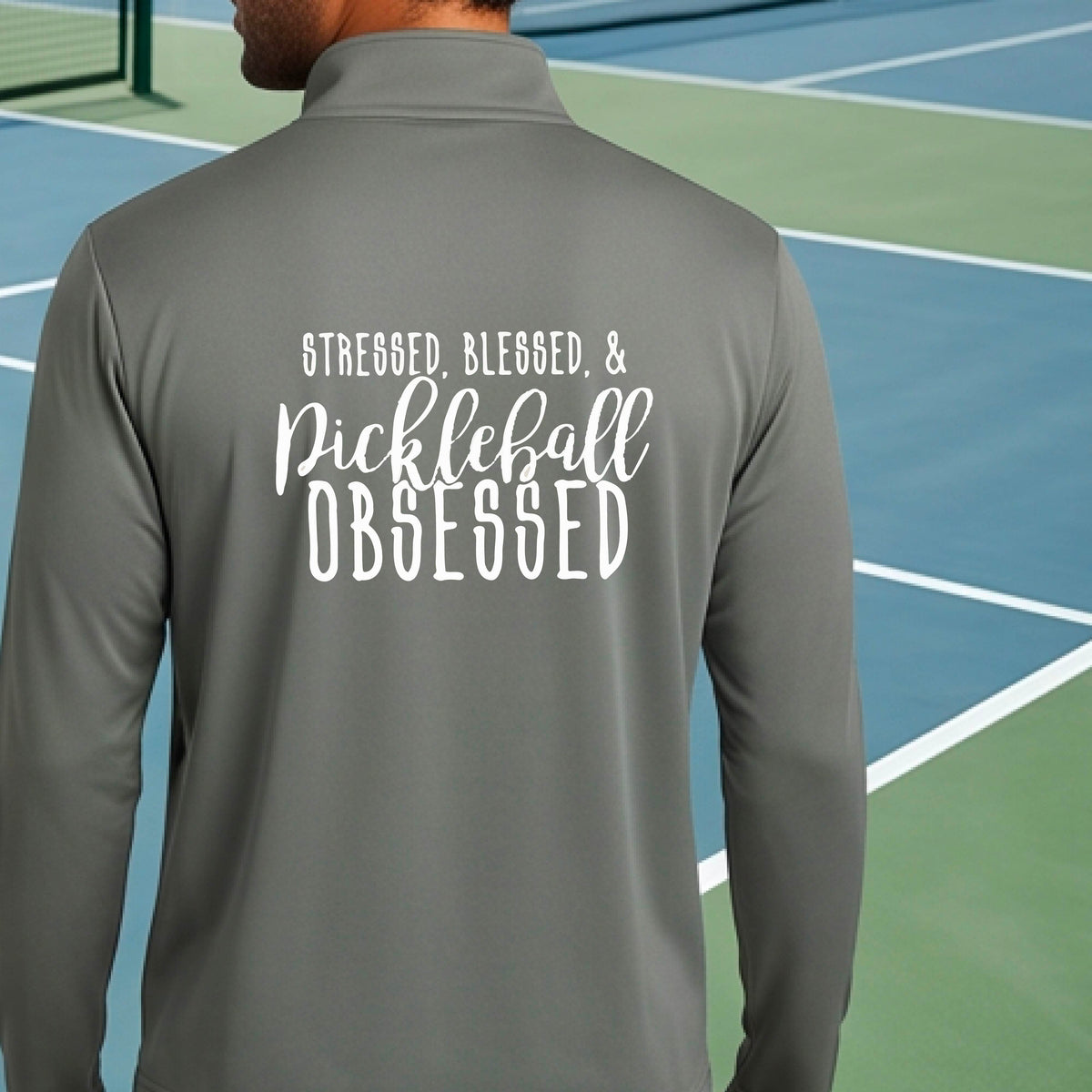 A man seen from behind on a pickleball court wearing a gray quarter-zip shirt with a white graphic of the words “Stressed, Blessed & Pickleball Obsessed” on the back of the shirt.