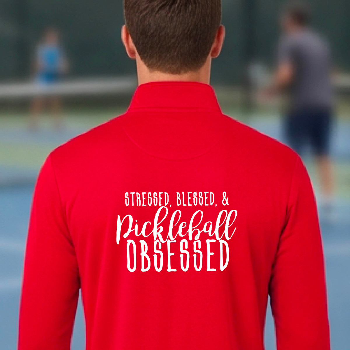 A man seen from behind on a pickleball court wearing a red quarter-zip shirt with a white graphic of the words “Stressed, Blessed & Pickleball Obsessed” on the back of the shirt.
