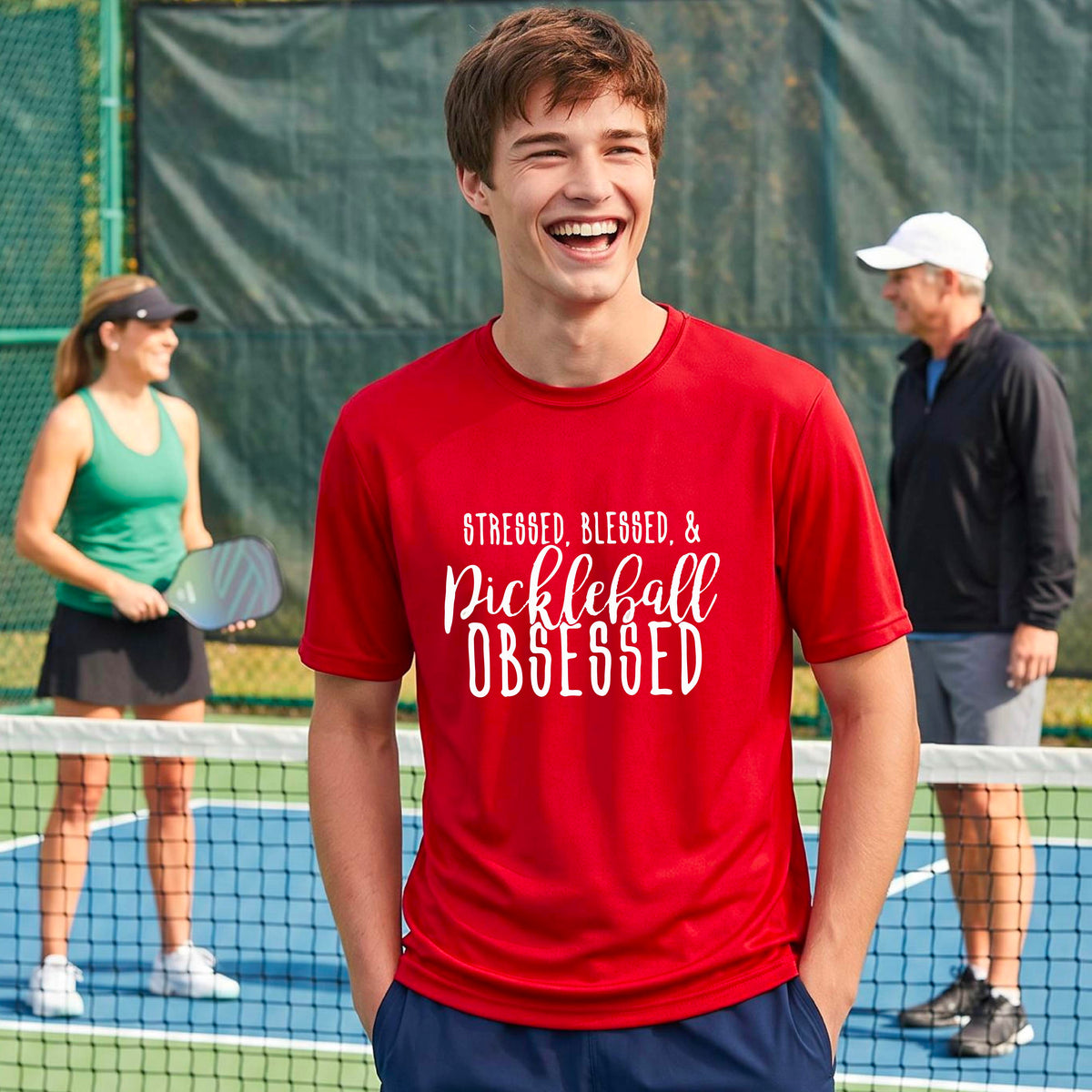 A man on a pickleball court wearing a red short-sleeve t-shirt with a white graphic of the words “Stressed, Blessed & Pickleball Obsessed” on the front of the shirt.
