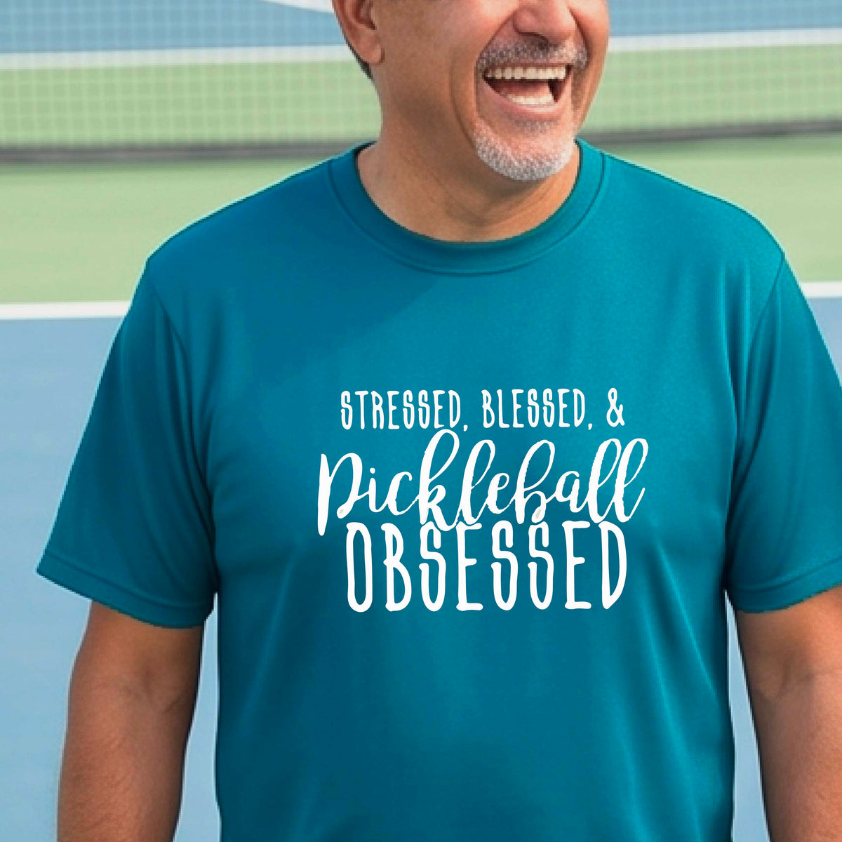A man on a pickleball court wearing a teal short-sleeve t-shirt with a white graphic of the words “Stressed, Blessed & Pickleball Obsessed” on the front of the shirt.