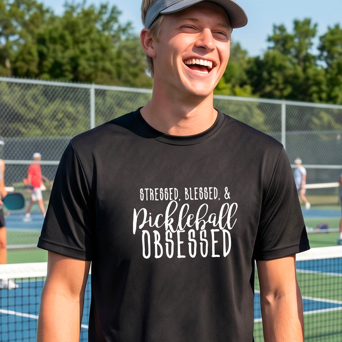 A man on a pickleball court wearing a black short-sleeve t-shirt with a white graphic of the words “Stressed, Blessed & Pickleball Obsessed” on the front of the shirt.