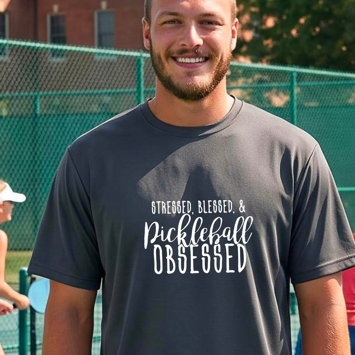 A man on a pickleball court wearing a gray short-sleeve t-shirt with a white graphic of the words “Stressed, Blessed & Pickleball Obsessed” on the front of the shirt.