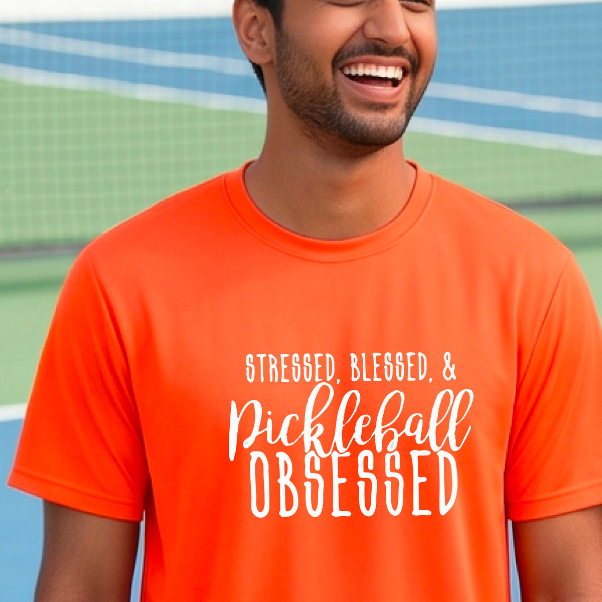 A man on a pickleball court wearing an orange short-sleeve t-shirt with a white graphic of the words “Stressed, Blessed & Pickleball Obsessed” on the front of the shirt.
