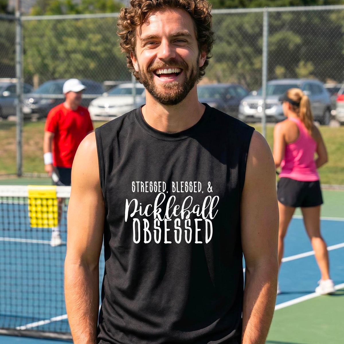A man on a pickleball court wearing a black sleeveless t-shirt with a white graphic of the words “Stressed, Blessed & Pickleball Obsessed” on the front of the shirt.