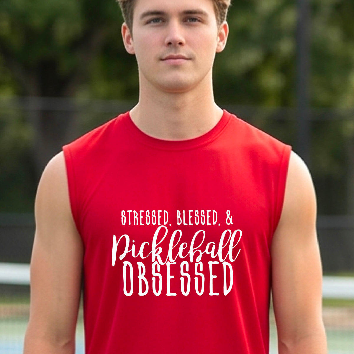 A man on a pickleball court wearing a red sleeveless t-shirt with a white graphic of the words “Stressed, Blessed & Pickleball Obsessed” on the front of the shirt.