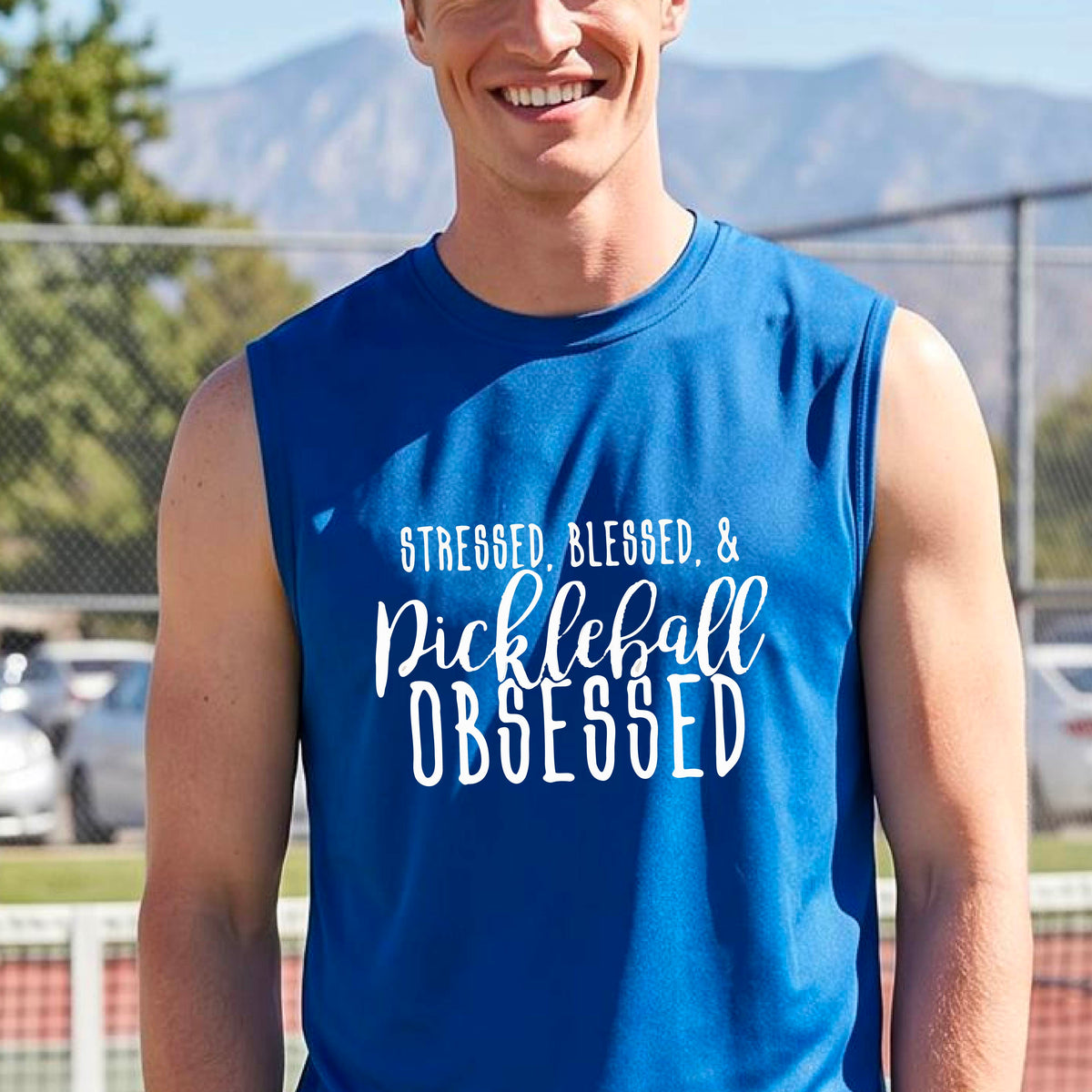 A man on a pickleball court wearing a royal blue sleeveless t-shirt with a white graphic of the words “Stressed, Blessed & Pickleball Obsessed” on the front of the shirt.