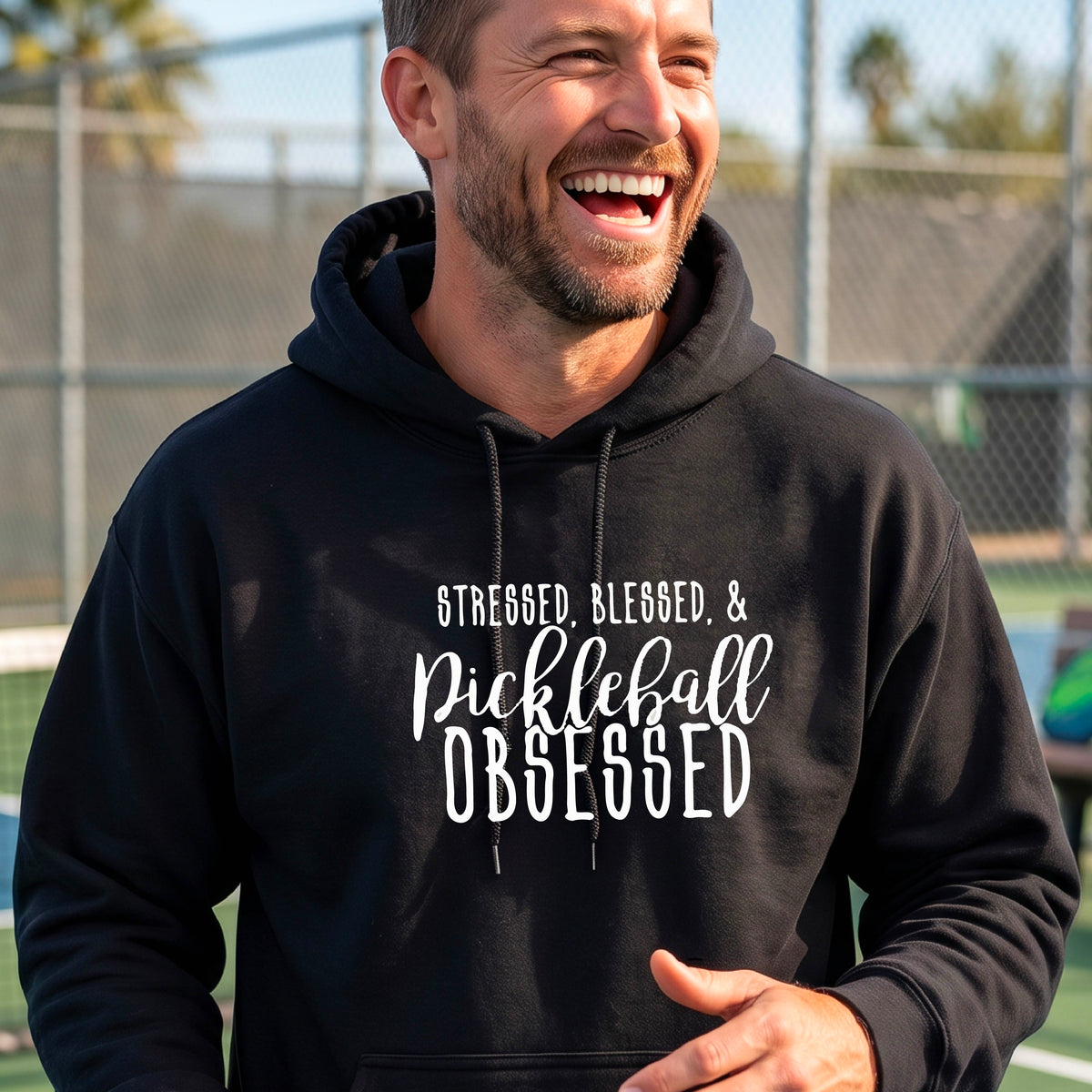 A man on a pickleball court wearing a black hoodie pullover with a white graphic of the words “Stressed, Blessed & Pickleball Obsessed” on the front of the shirt.