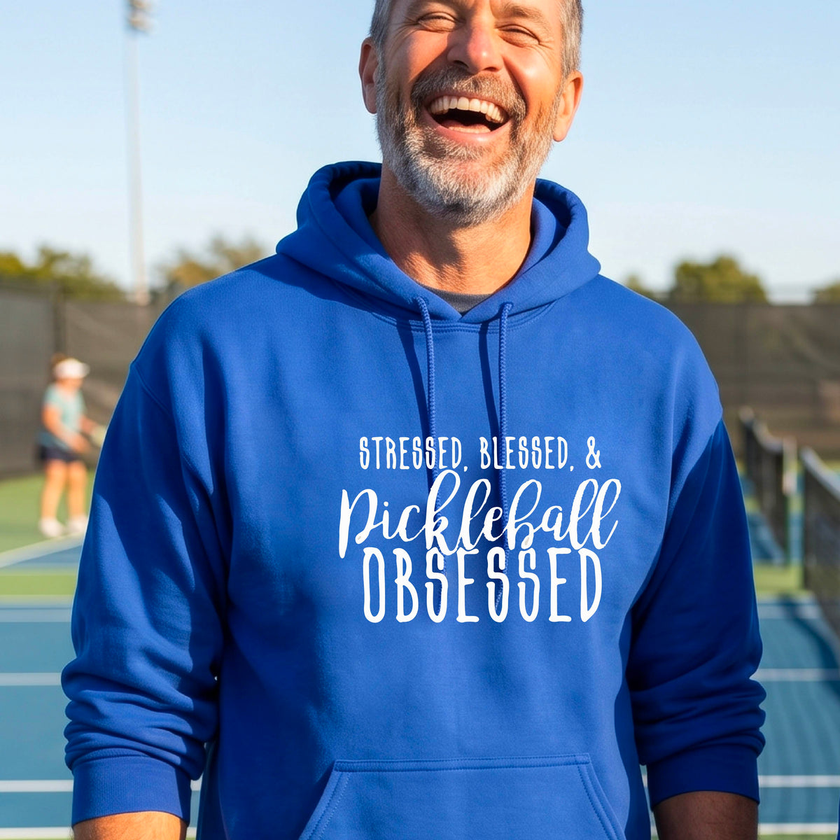 A man on a pickleball court wearing a royal blue hoodie pullover with a white graphic of the words “Stressed, Blessed & Pickleball Obsessed” on the front of the shirt.