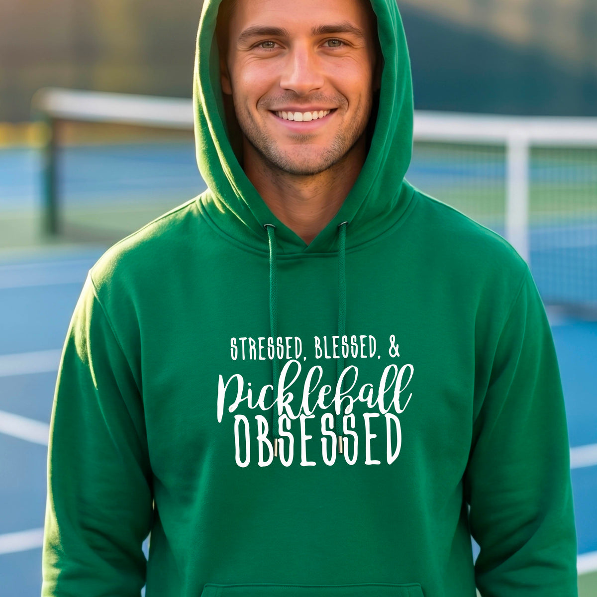 A man on a pickleball court wearing a green hoodie pullover with a white graphic of the words “Stressed, Blessed & Pickleball Obsessed” on the front of the shirt.