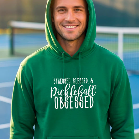 A man on a pickleball court wearing a green hoodie pullover with a white graphic of the words “Stressed, Blessed & Pickleball Obsessed” on the front of the shirt.