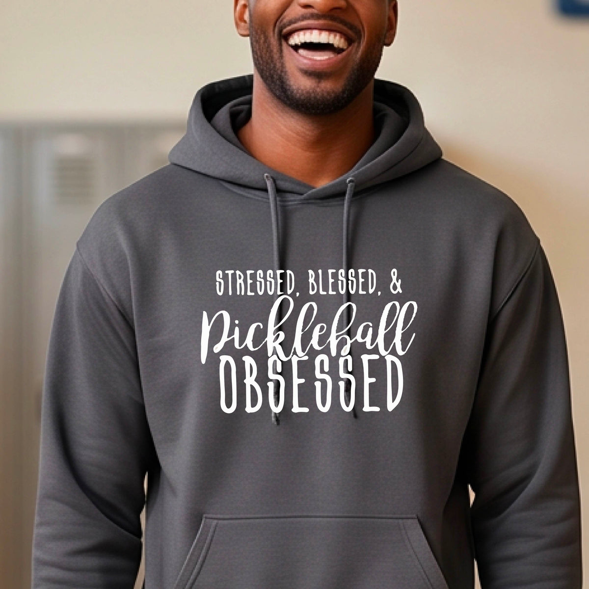 A man on a pickleball court wearing a gray hoodie pullover with a white graphic of the words “Stressed, Blessed & Pickleball Obsessed” on the front of the shirt.