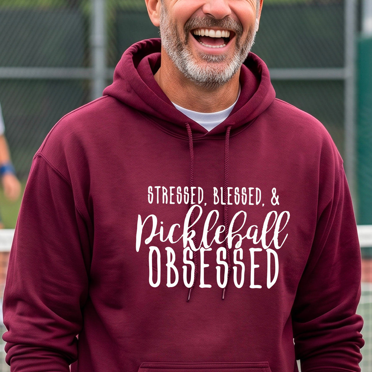 A man on a pickleball court wearing a maroon hoodie pullover with a white graphic of the words “Stressed, Blessed & Pickleball Obsessed” on the front of the shirt.