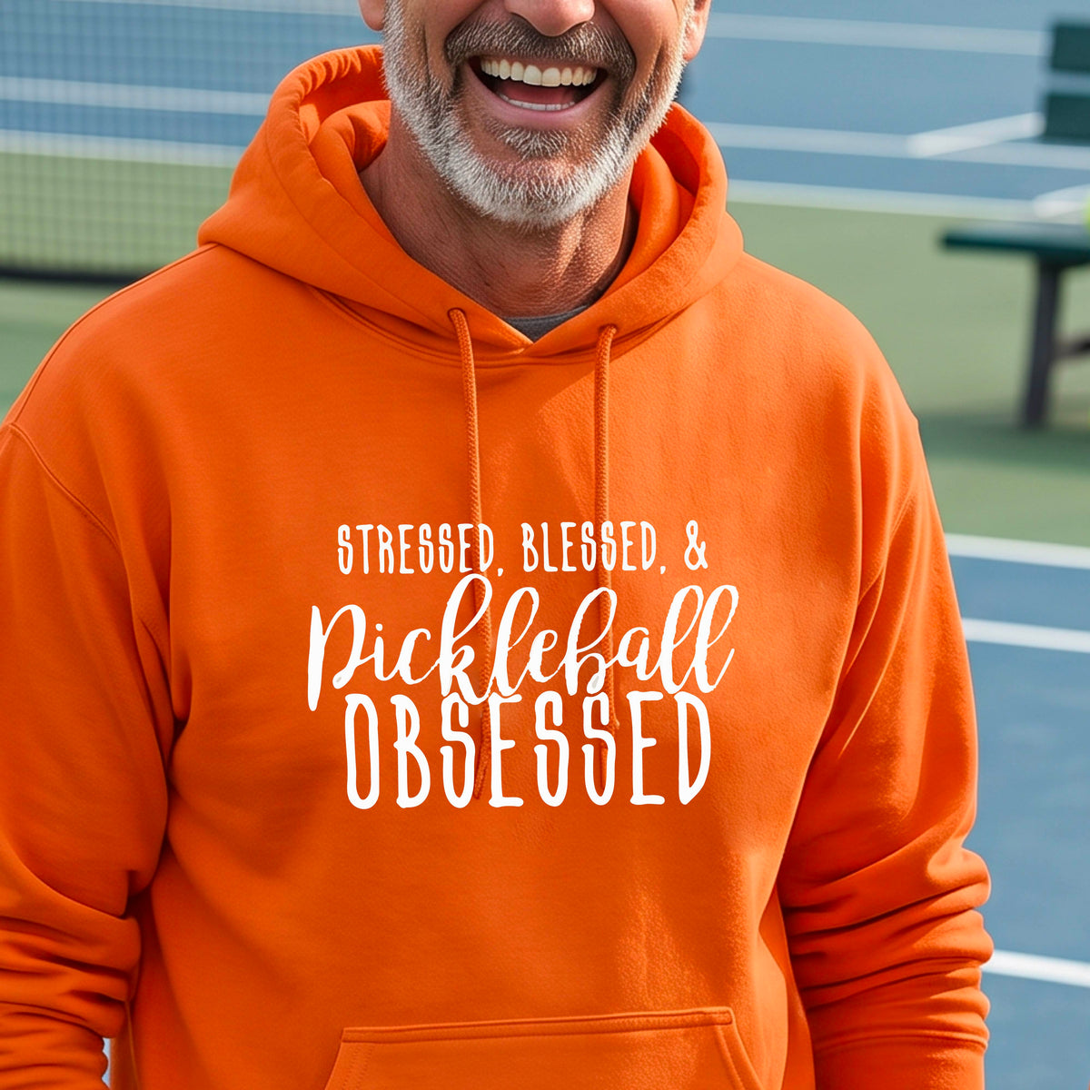 A man on a pickleball court wearing an orange hoodie pullover with a white graphic of the words “Stressed, Blessed & Pickleball Obsessed” on the front of the shirt.
