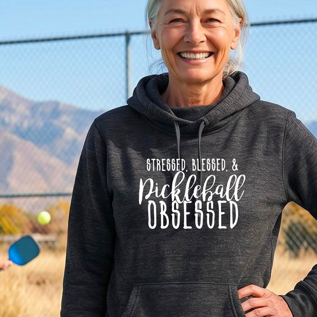 A woman on a pickleball court wearing a black hoodie pullover with a white graphic of the words “Stressed, Blessed & Pickleball Obsessed” on the front of the shirt.