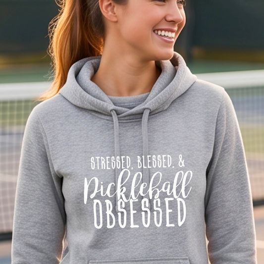 A woman on a pickleball court wearing a gray hoodie pullover with a white graphic of the words “Stressed, Blessed & Pickleball Obsessed” on the front of the shirt.