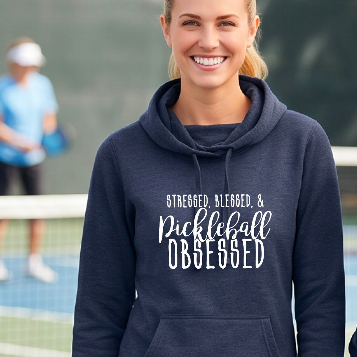 A woman on a pickleball court wearing a navy blue hoodie pullover with a white graphic of the words “Stressed, Blessed & Pickleball Obsessed” on the front of the shirt.