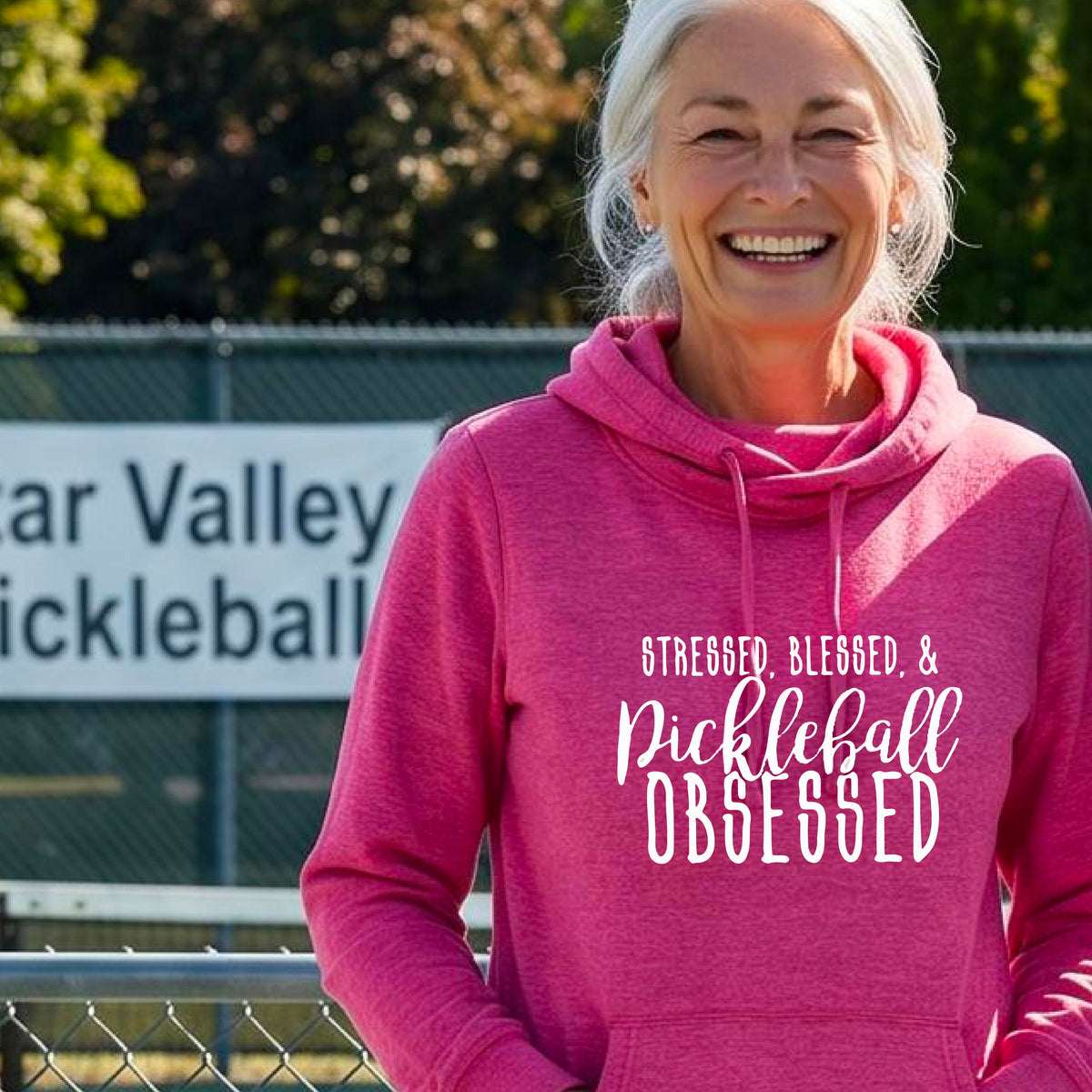 A woman on a pickleball court wearing a pink hoodie pullover with a white graphic of the words “Stressed, Blessed & Pickleball Obsessed” on the front of the shirt.