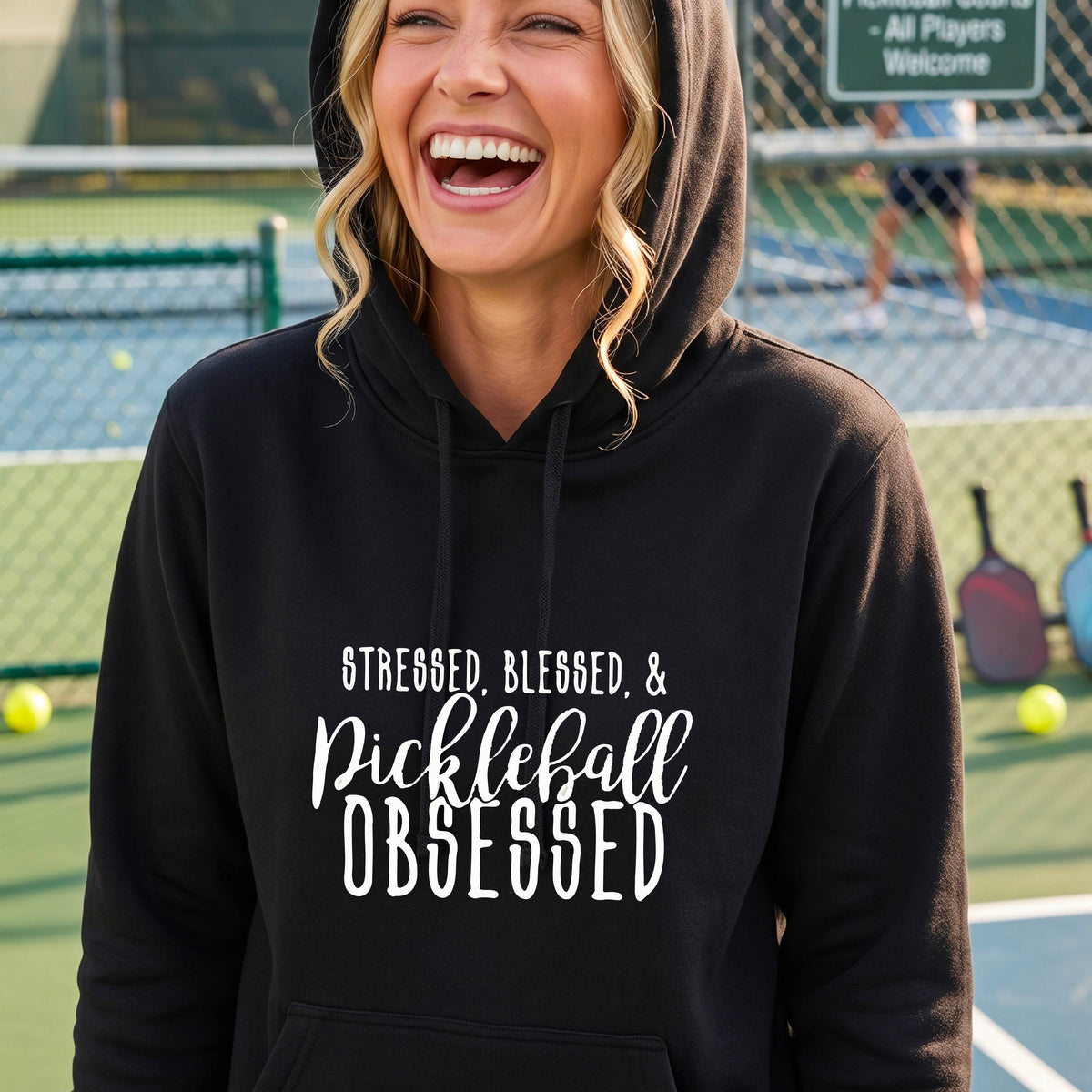 A woman wearing a black fitted hoodie pullover with a white graphic of the words “Stressed, Blessed & Pickleball Obsessed” on the front of the hoodie.