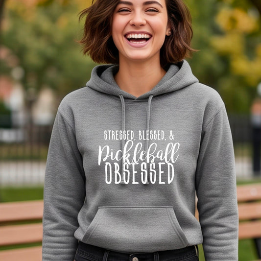 A woman wearing a gray fitted hoodie pullover with a white graphic of the words “Stressed, Blessed & Pickleball Obsessed” on the front of the hoodie.