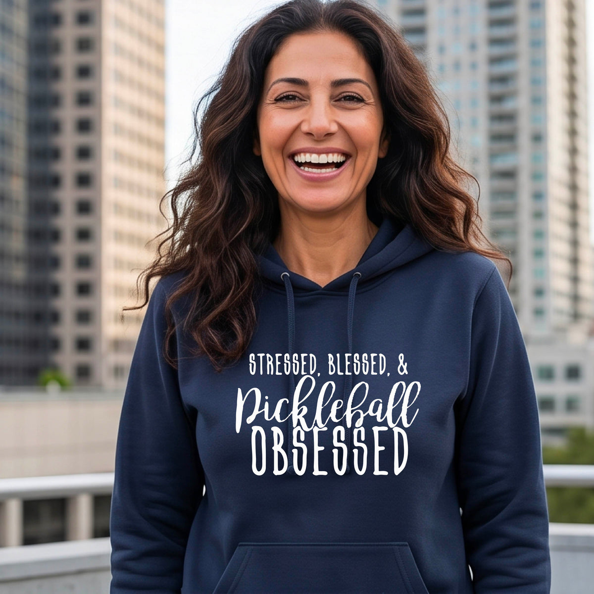 A woman wearing a navy blue fitted hoodie pullover with a white graphic of the words “Stressed, Blessed & Pickleball Obsessed” on the front of the hoodie.