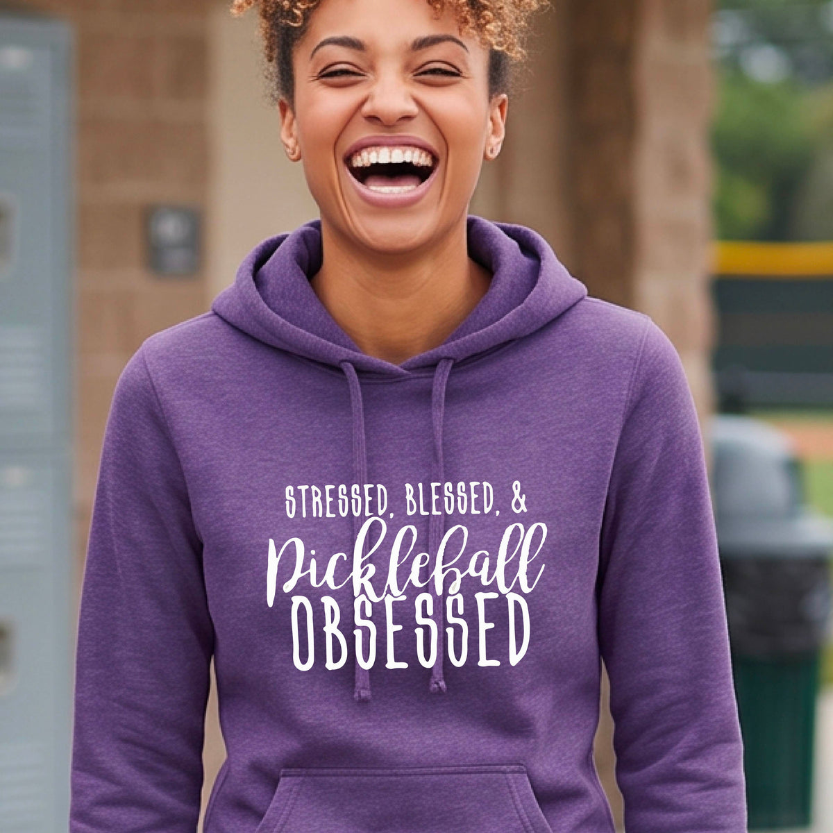 A woman wearing a purple fitted hoodie pullover with a white graphic of the words “Stressed, Blessed & Pickleball Obsessed” on the front of the hoodie.