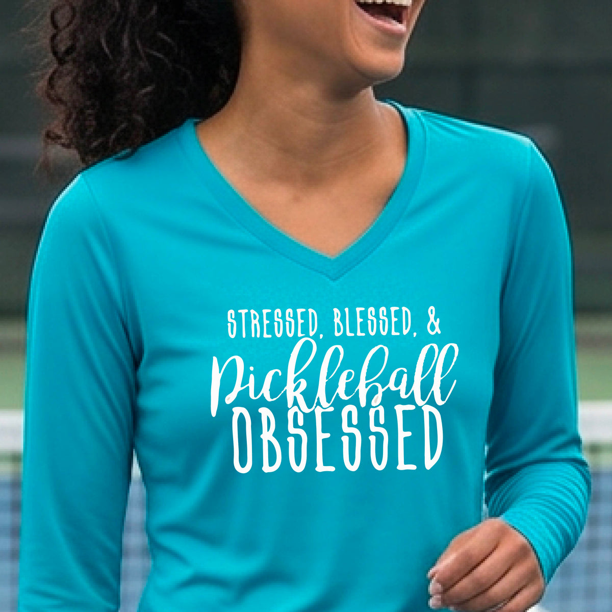 A woman on a pickleball court wearing a bright blue hoodie pullover with a white graphic of the words “Stressed, Blessed & Pickleball Obsessed” on the front of the shirt.