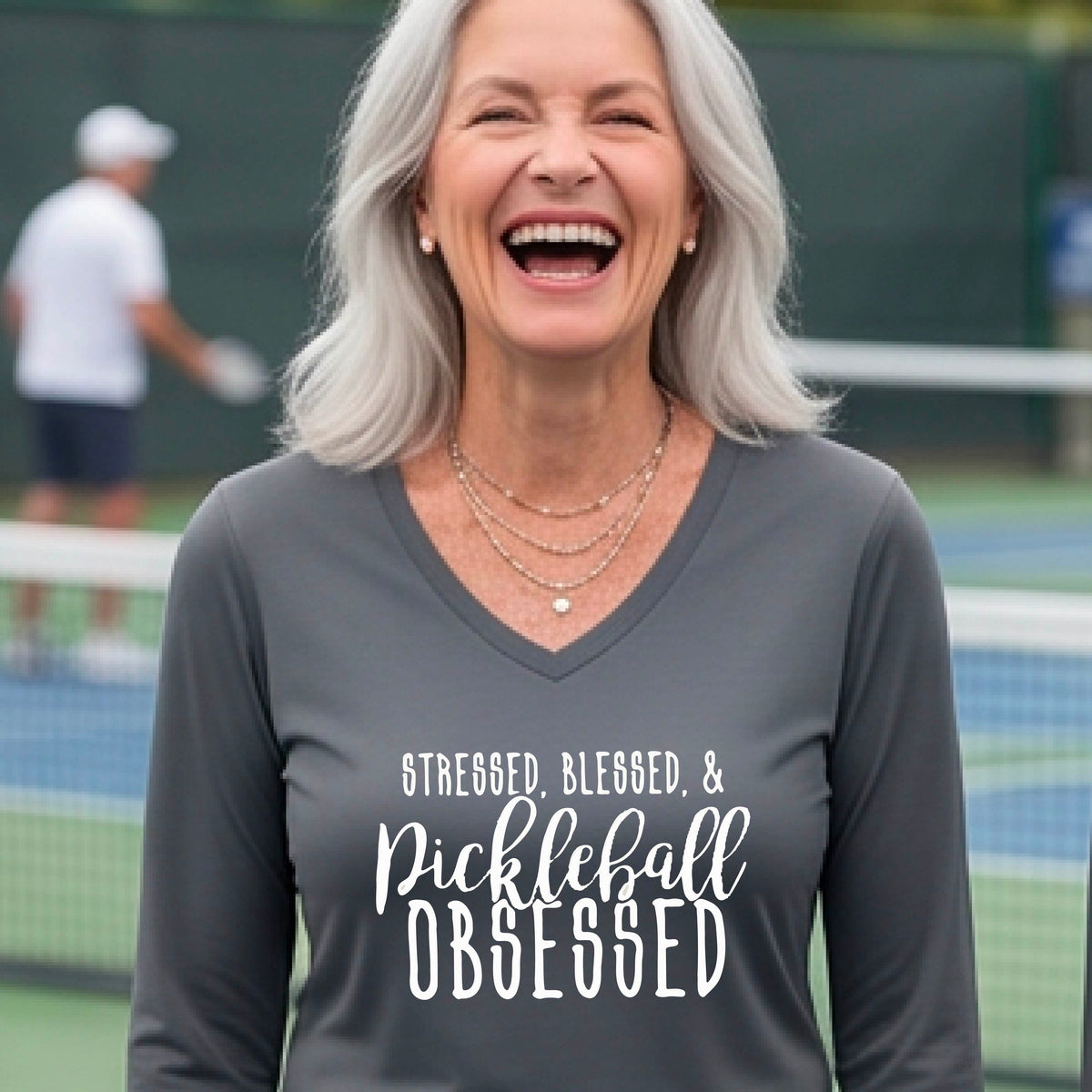 A woman on a pickleball court wearing a gray hoodie pullover with a white graphic of the words “Stressed, Blessed & Pickleball Obsessed” on the front of the shirt.