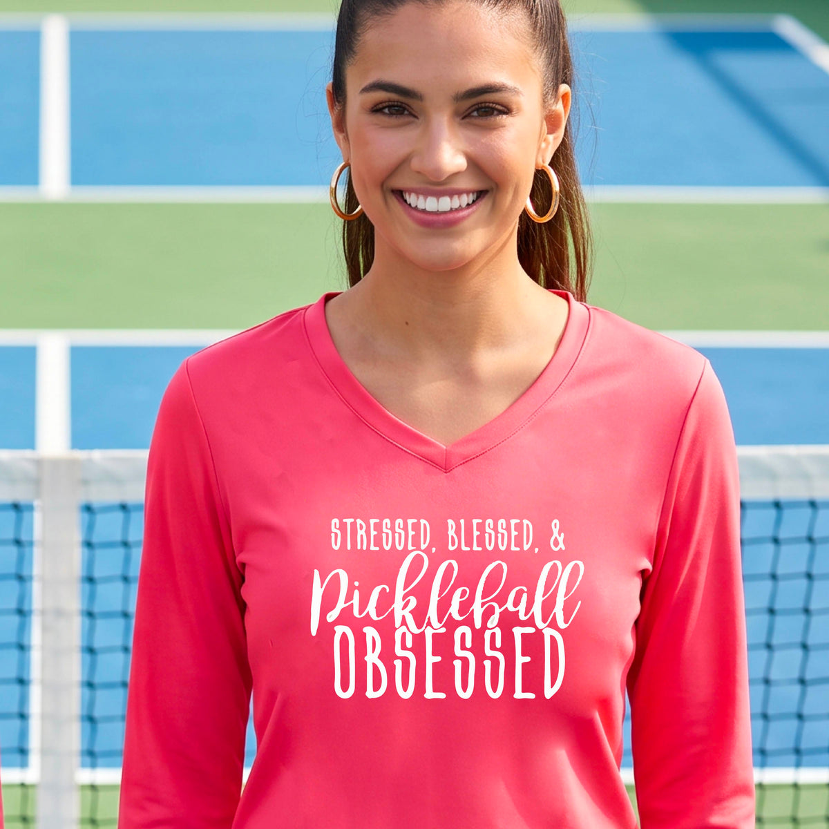 A woman on a pickleball court wearing a coral pink hoodie pullover with a white graphic of the words “Stressed, Blessed & Pickleball Obsessed” on the front of the shirt.