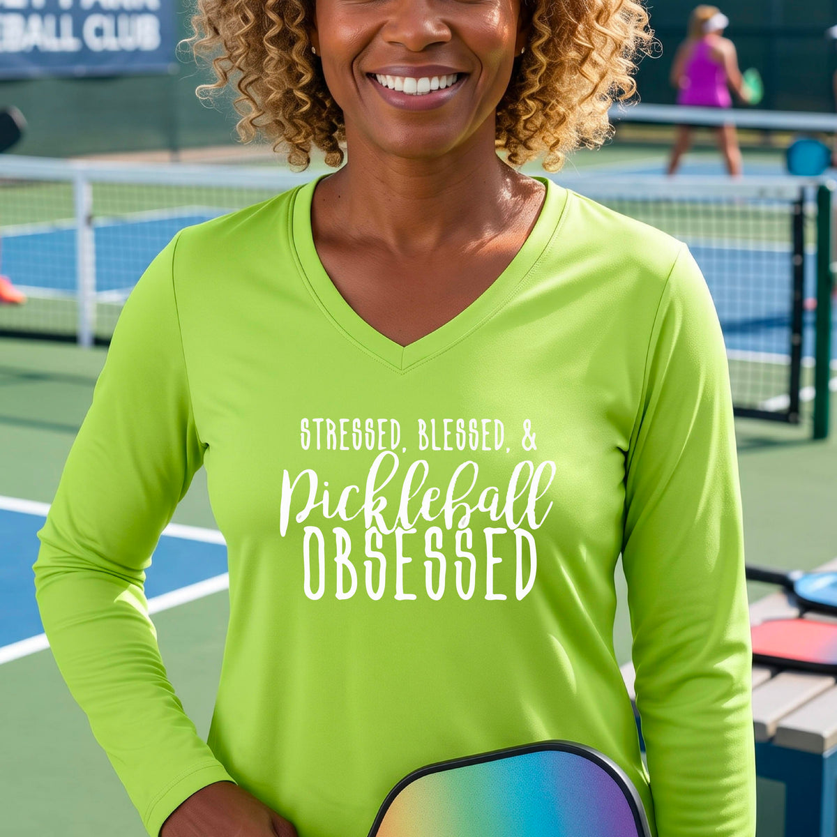 A woman on a pickleball court wearing a lime green hoodie pullover with a white graphic of the words “Stressed, Blessed & Pickleball Obsessed” on the front of the shirt.