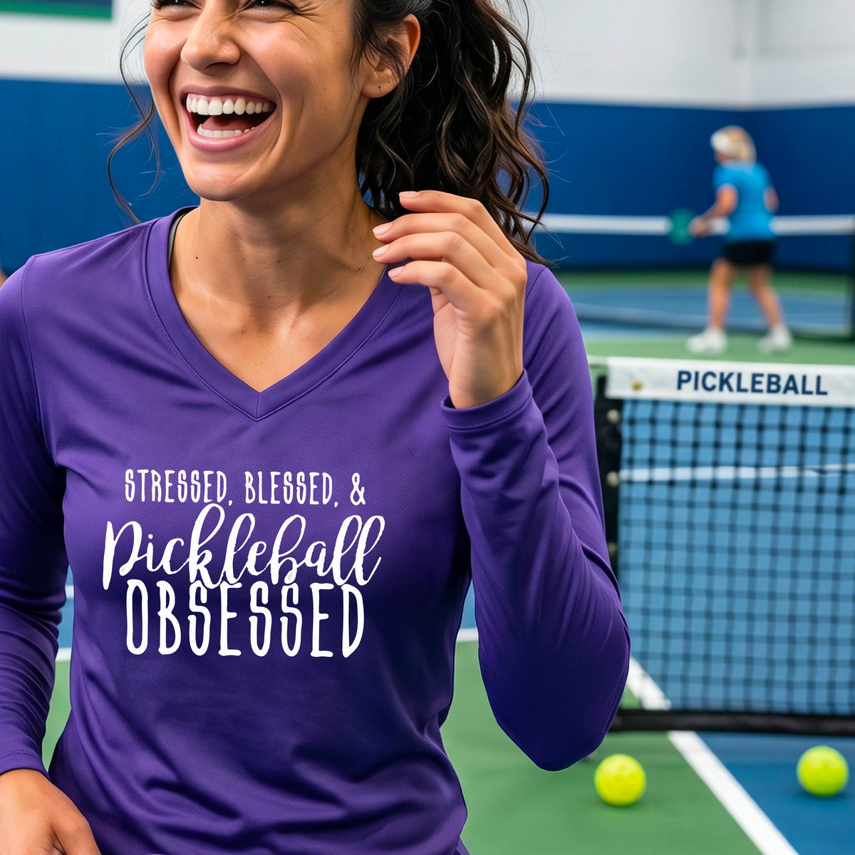 A woman on a pickleball court wearing a purple hoodie pullover with a white graphic of the words “Stressed, Blessed & Pickleball Obsessed” on the front of the shirt.